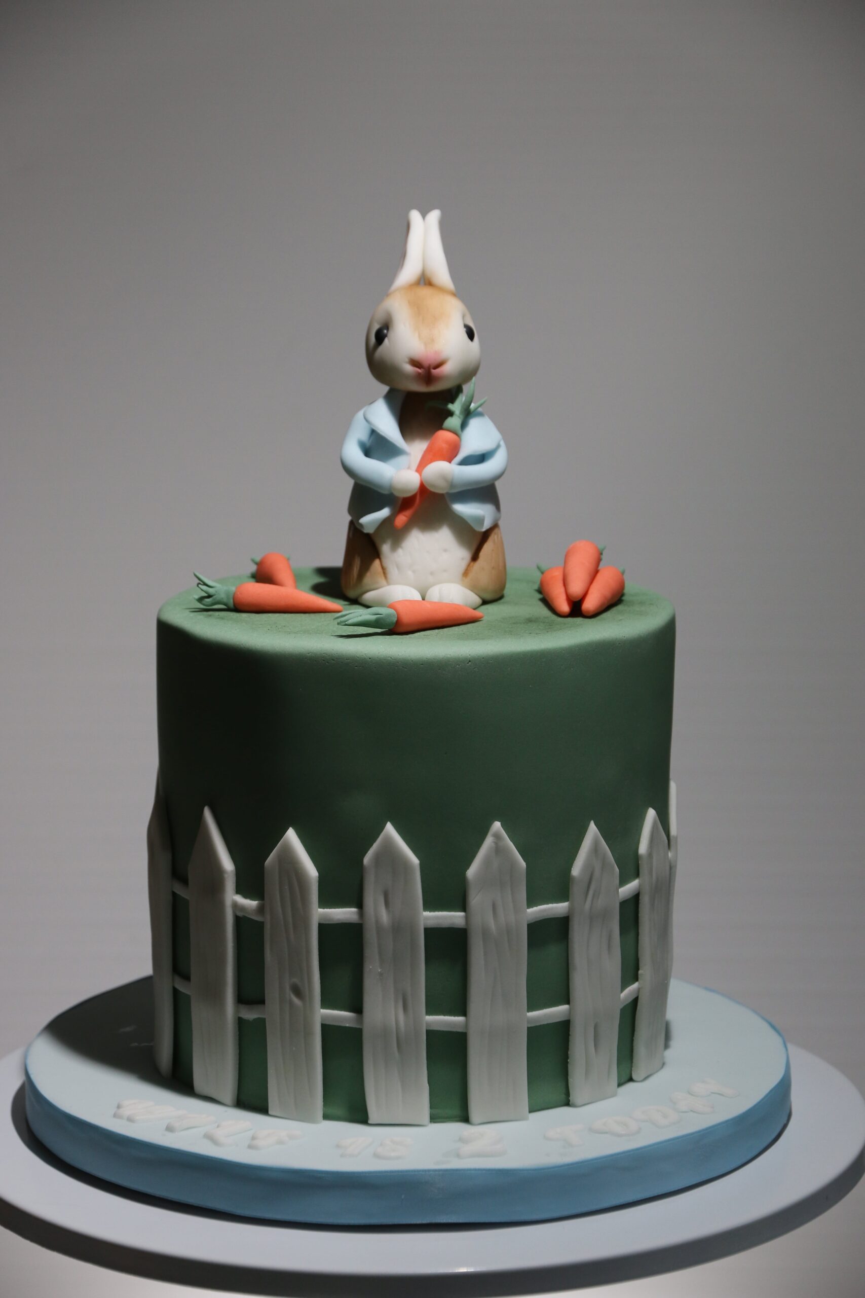 Peter Rabbit eating carrots on top of a green coloured cake surrounded by a picket fence